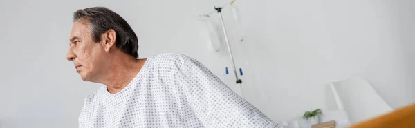 Vista laterale di uomo anziano in abito paziente guardando altrove nel reparto ospedaliero, banner — Foto stock