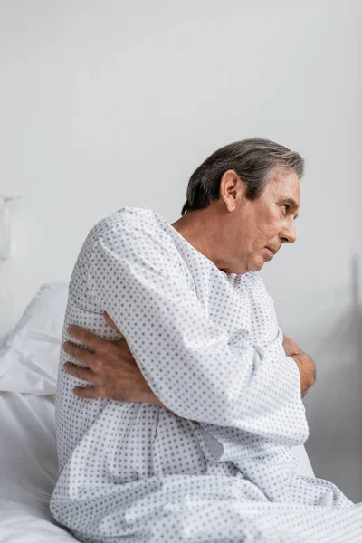 Side view of sick senior man sitting on bed in hospital ward — Stock Photo