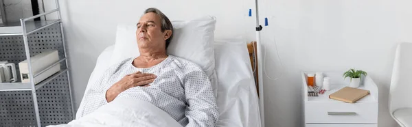 Elderly man in patient gown lying near intravenous therapy and pills in clinic, banner — Stock Photo