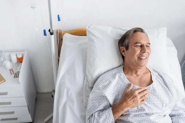 Высокий угол обзора улыбающегося пожилого человека в больничном халате лежащего на кровати в больничной палате — стоковое фото