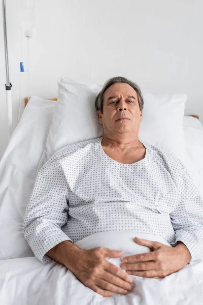 Sick grey haired patient in gown looking at camera while lying on bed in clinic - foto de stock