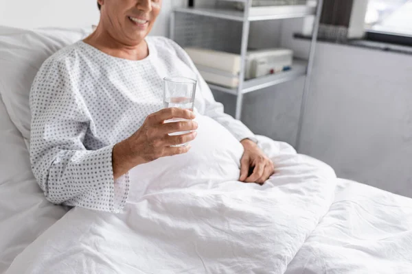 Cropped view of blurred patient holding glass of water while sitting on hospital bed - foto de stock