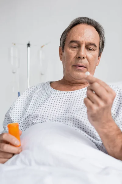 Grey haired patient holding pill while sitting on hospital bed - foto de stock