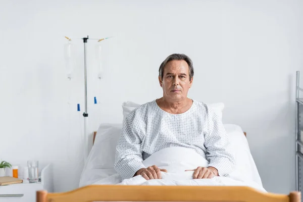 Paciente sênior olhando para a câmera enquanto estava sentado na cama na enfermaria do hospital — Fotografia de Stock
