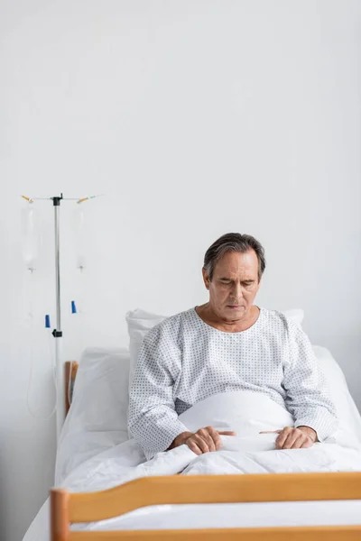 Paciente idoso perturbado de vestido sentado na cama perto de terapia intravenosa na clínica — Fotografia de Stock