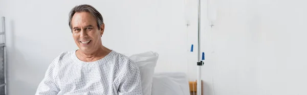 Senior patient smiling at camera near intravenous therapy in clinic, banner — Stock Photo