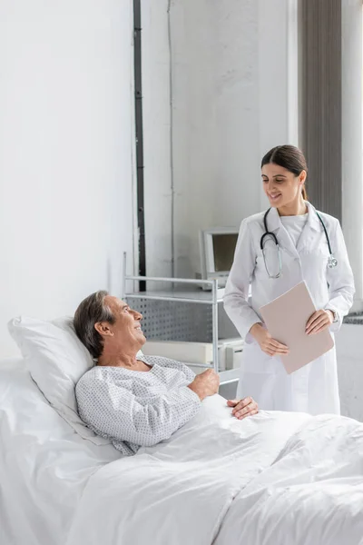 Улыбающийся доктор держит бумажную папку рядом с больным пациентом в клинике — стоковое фото