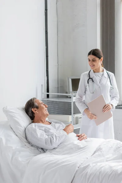 Smiling doctor holding paper folder near sick patient in clinic — Stock Photo