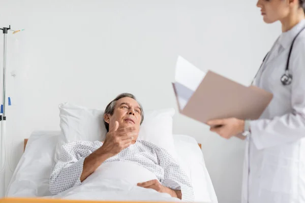 Elderly patient talking to blurred doctor with paper folder in hospital ward — Stock Photo