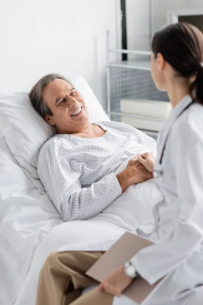 Medico sfocato che tiene per mano un paziente anziano sorridente in clinica — Foto stock
