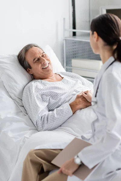 Blurred doctor holding hand of smiling senior patient in clinic — Stock Photo