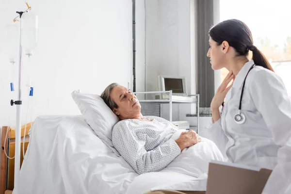 Doctor with paper folder touching neck while talking to senior patient in hospital ward - foto de stock