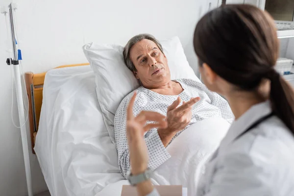 Senior patient talking to blurred doctor with paper folder in hospital ward - foto de stock