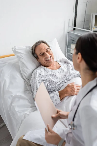 Paziente anziano positivo che guarda il medico sfocato con cartella cartacea nel reparto ospedaliero — Foto stock