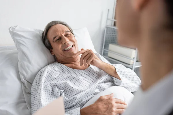 Cheerful elderly patient looking at blurred doctor in hospital ward — Stock Photo