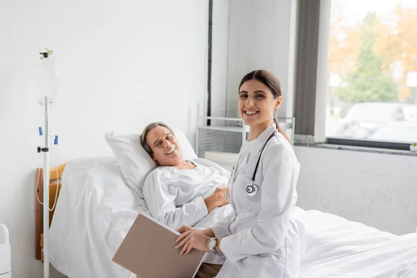 Positive doctor with paper folder looking at camera near senior patient in hospital ward — Stock Photo