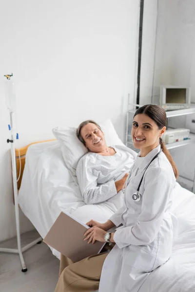 Positive doctor holding paper folder and looking at camera near blurred senior patient in clinic - foto de stock