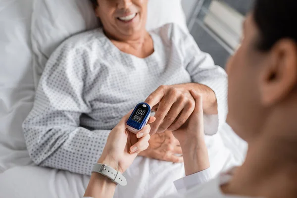 Blurred doctor wearing pulse oximeter on finger of smiling patient in hospital ward - foto de stock