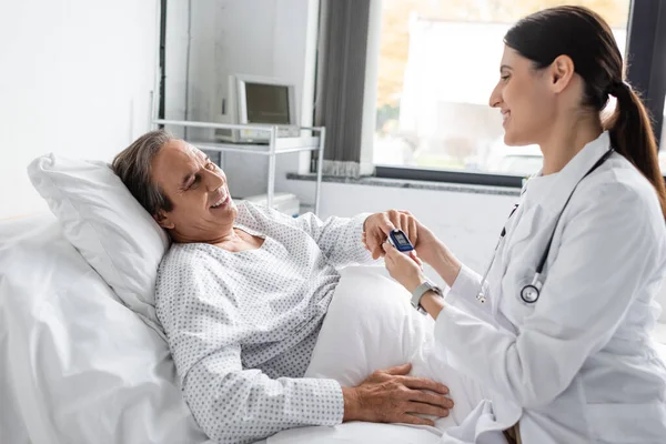 Positive doctor wearing pulse oximeter on finger of senior patient in hospital ward — Stock Photo