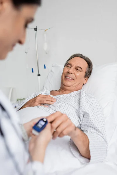 Lächelnde Seniorin mit Pulsoximeter blickt verschwommenen Arzt in Klinik an — Stockfoto