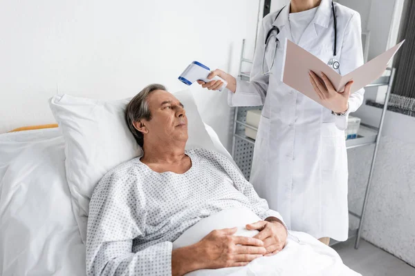 Врач с бумажной папкой держит пирометр рядом со старшим пациентом в больнице — стоковое фото