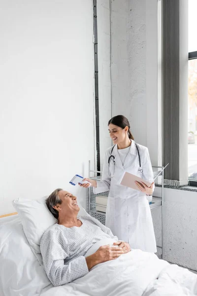 Cheerful doctor with paper folder holding pyrometer near senior patient in hospital ward — Photo de stock