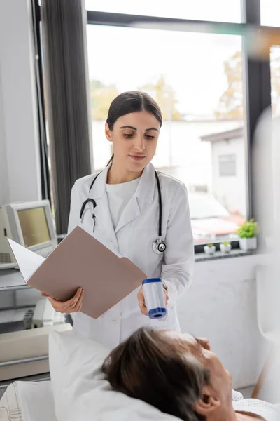 Brunette doctor with paper folder checking temperature of senior patient in hospital ward — Stock Photo