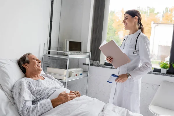 Positive elderly patient looking at doctor with paper folder and pyrometer in hospital ward — Stock Photo