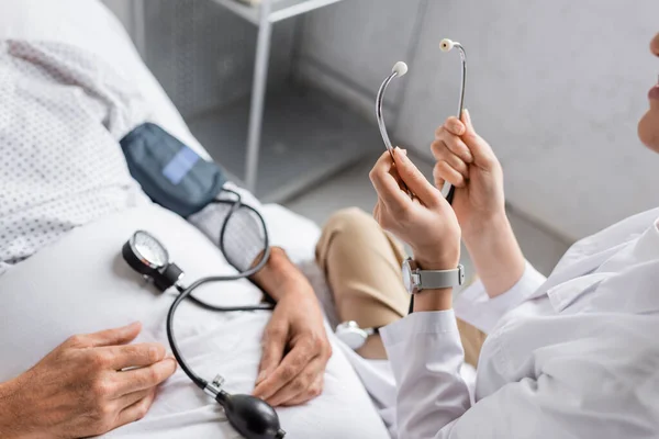 Cropped view of doctor holding stethoscope near tonometer and sick patient on hospital bed — Stock Photo