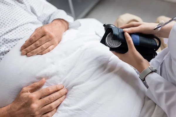 Обрезанный вид врача, держащего тонометр рядом с больным пациентом на больничной койке — стоковое фото