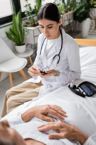 Doctor holding glucometer near senior patient on bed in clinic — Stock Photo