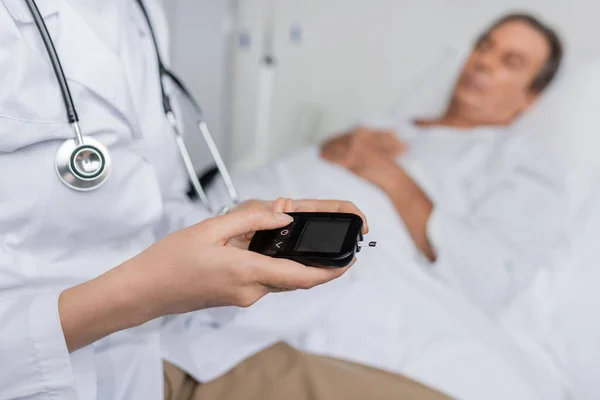 Doctor holding glucometer near blurred patient on hospital bed - foto de stock