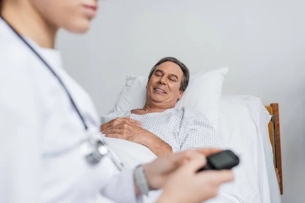 Smiling elderly patient looking at blurred doctor with glucometer in hospital ward — Stock Photo