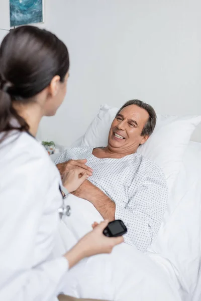 Smiling senior patient looking at blurred doctor with glucometer in clinic — Stock Photo