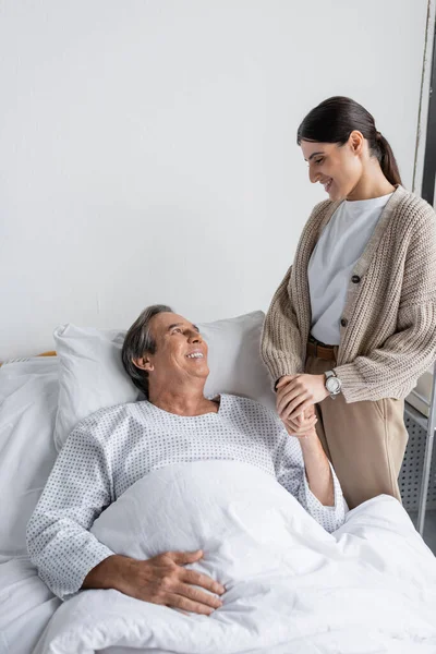 Smiling daughter holding hand of father lying on bed in hospital — Stock Photo