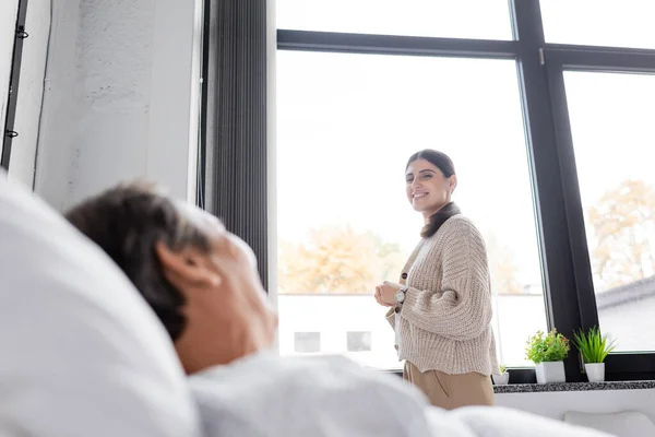 Smiling woman looking at blurred father lying on hospital bed — Stock Photo