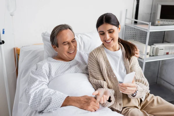 Улыбающаяся женщина держит смартфон рядом со старшим отцом на больничной койке — стоковое фото