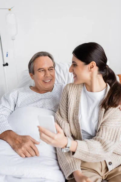 Старший пациент смотрит на улыбающуюся дочь со смартфоном в палате больницы — стоковое фото