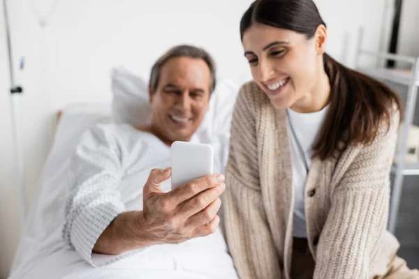Blurred senior patient holding smartphone near daughter in hospital ward - foto de stock