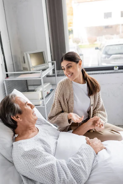Positive woman holding hand of father in patient gown and talking in hospital ward — Stock Photo