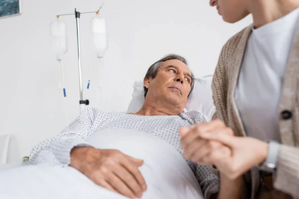Displeased elderly man holding hand of blurred daughter while lying on bed in clinic — Stock Photo