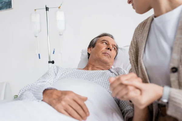 Dispiaciuto uomo anziano che tiene la mano della figlia offuscata mentre sdraiato sul letto in clinica — Foto stock