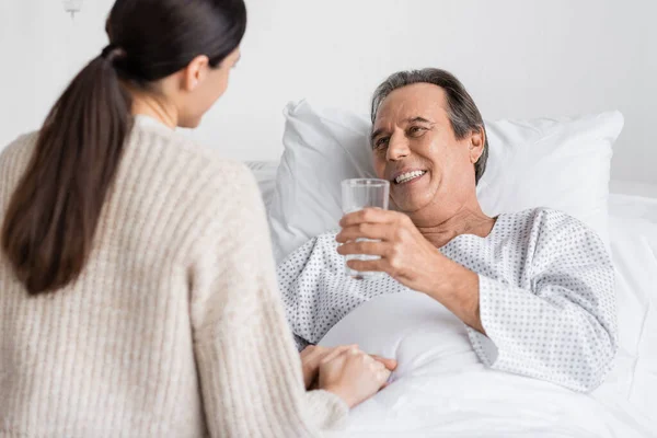 Sorridente paziente anziano che tiene un bicchiere d'acqua vicino alla figlia offuscata in clinica — Foto stock