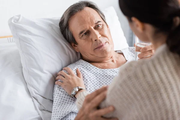 Paziente anziano che tiene un bicchiere d'acqua e guarda la figlia sfocata nel reparto ospedaliero — Foto stock
