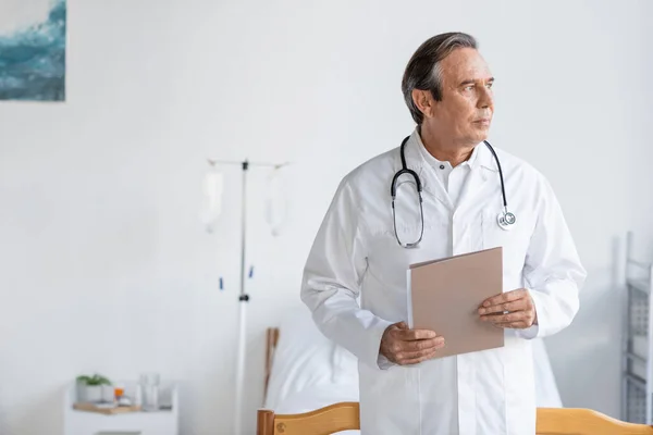 Elderly doctor holding paper folder in hospital ward — Stock Photo