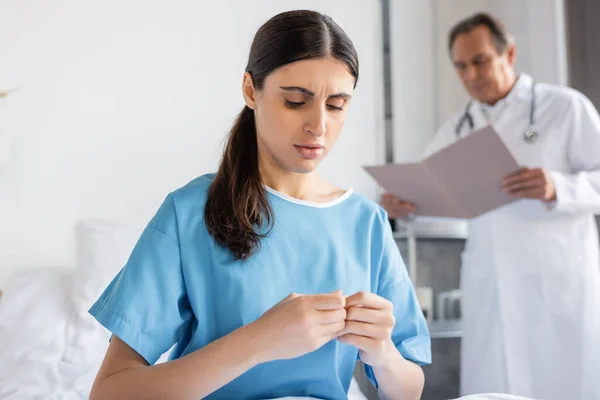 Nervous patient in gown sitting on bed near blurred doctor in hospital ward — Stock Photo