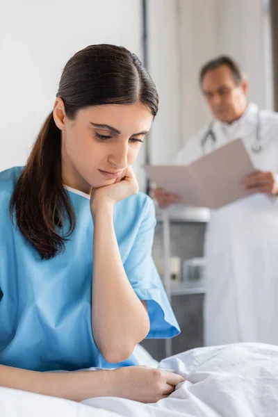 Upset patient sitting on bed near blurred doctor in hospital — Stock Photo