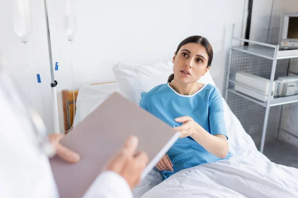 Brunette patient talking to blurred doctor with paper folder in hospital ward — Stock Photo