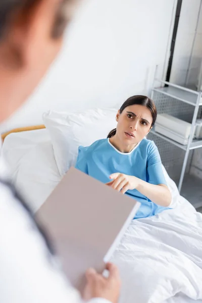 Paziente preoccupato in abito parlando con medico sfocato con cartella cartacea in ospedale — Foto stock