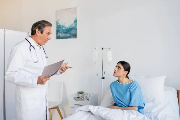 Senior doctor with paper folder talking to brunette patient in hospital ward - foto de stock